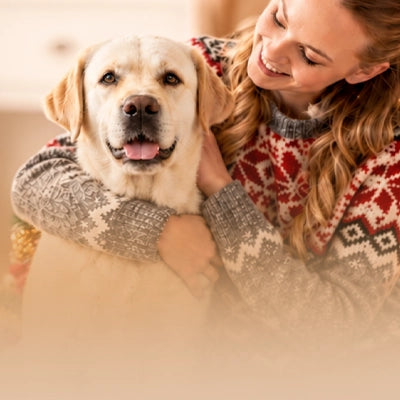 Fall Pet Catalog 2026. A woman in a red and gray patterned knit sweater hugs a light cream-colored Labrador Retriever in a warm, softly lit indoor setting.
